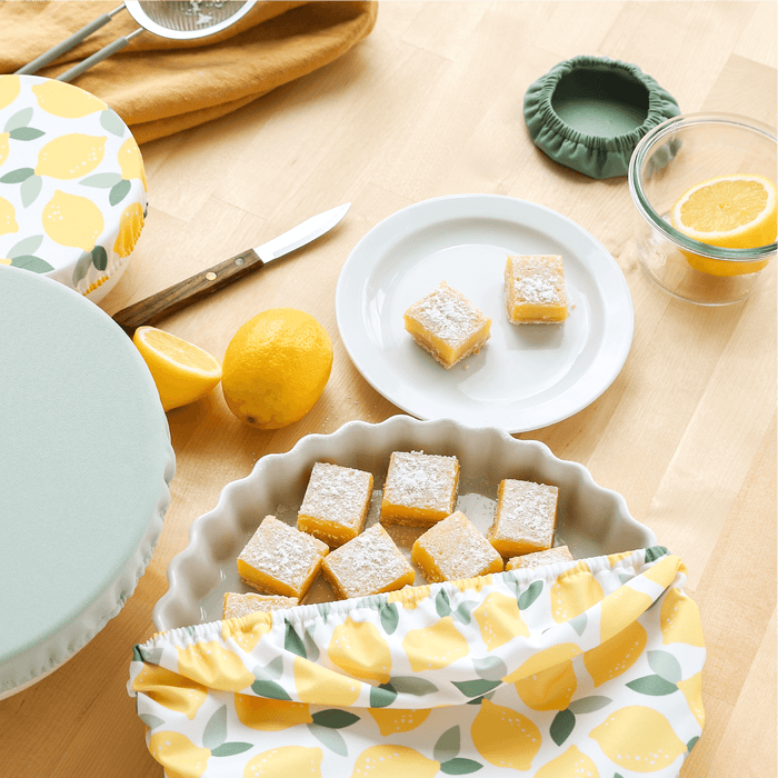Lemon bars on a table with a lemon-themed Bowl Caps, green Bowl Caps, and lemons.