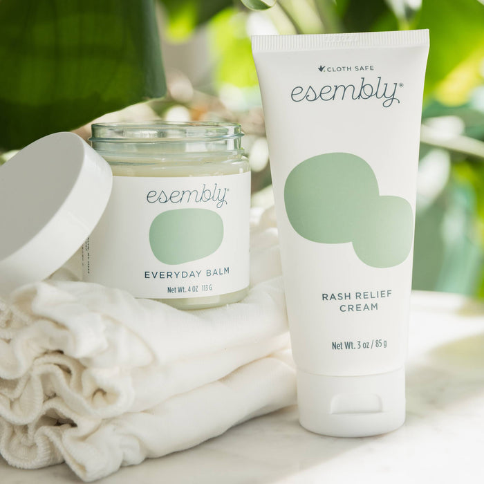 An Esembly Rash Relief Cream next to an Esembly Everyday Balm sitting on a stack of two Esembly Inners in front of leaves