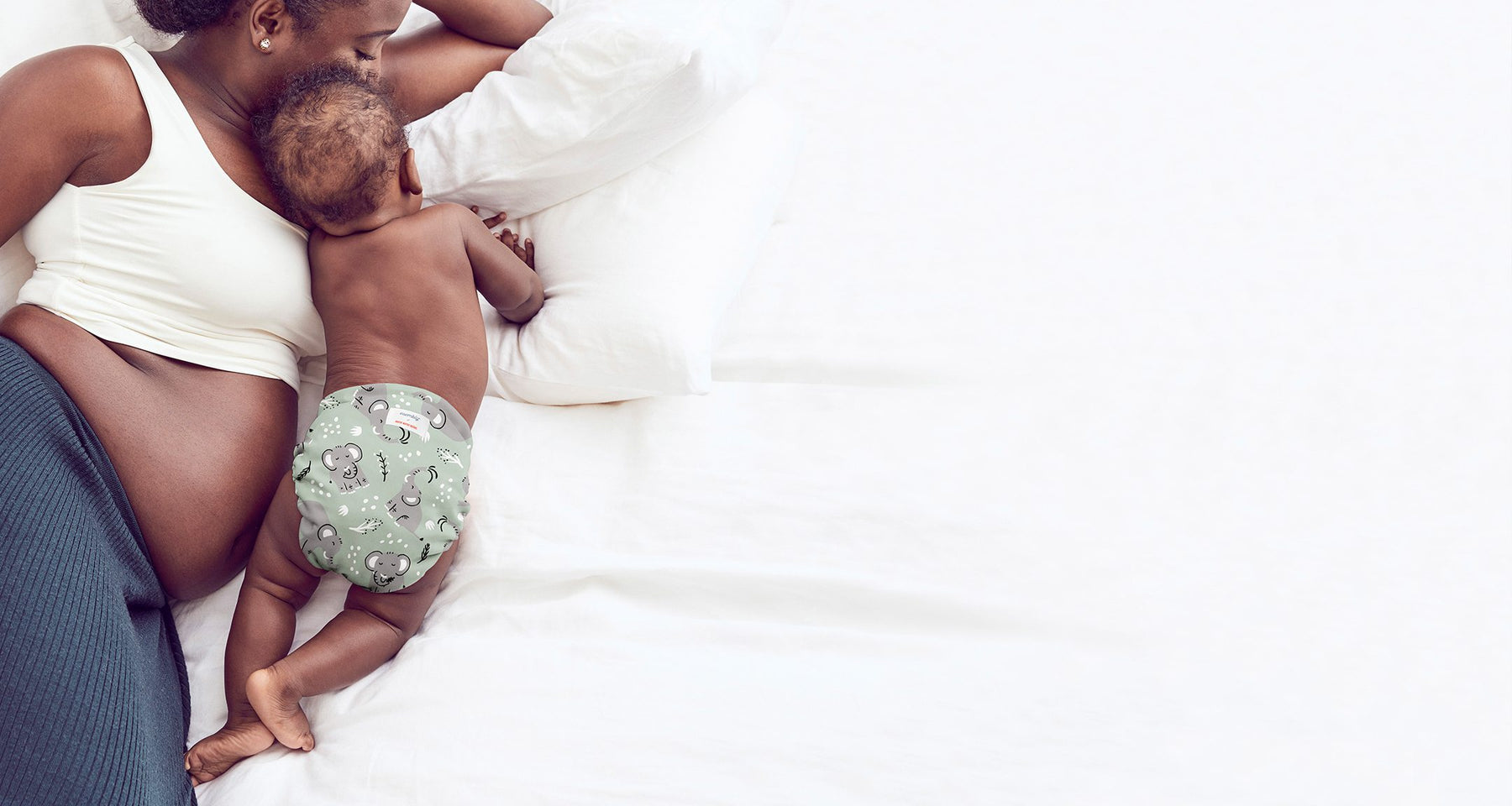 Back-view of baby wearing an Elephants Outer (a light green background with light grey elephants and white and black tree limbs and white sporadic polkadots ), crawling next to a woman wearing a grayish blue skirt with a white bra, laying on her side on white bed and pillows