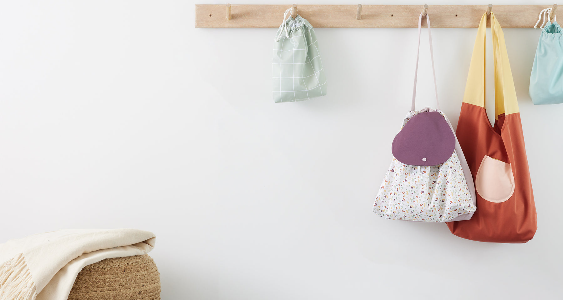 Wooden pegs with ditty bags (in Mist and Lattice (a light green background with white lined grid pattern), Carryall Bag in Clay (red body, yellow strap, and pink pocket), and a Day Bag in Terrazzo (an off-white background with a terrazzo pattern in light purple, dark purple, light green, dark green, yellow, and clay speckles body and strap with a dark purple flap) hanging on it and a natural fiber poof with a blanket on top of it to its left