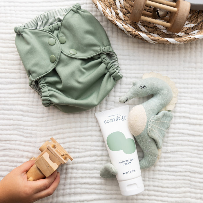 Rash Relief Cream (a white tube with a green splotch and dark blue text) nestled up against a light blue stuffed sea horse, next to a green diaper, a basket with wooden toy in it, and a child's hand holding a wooden toy