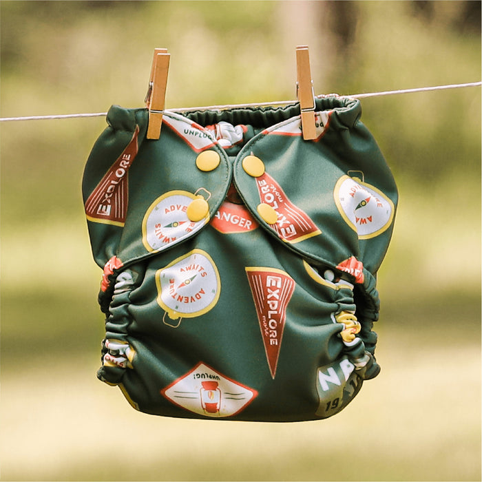An Adventure Badges Outer (a dark green background with camping and outdoorsy themed badge illustrations) hanging on a laundry line with clothing pins