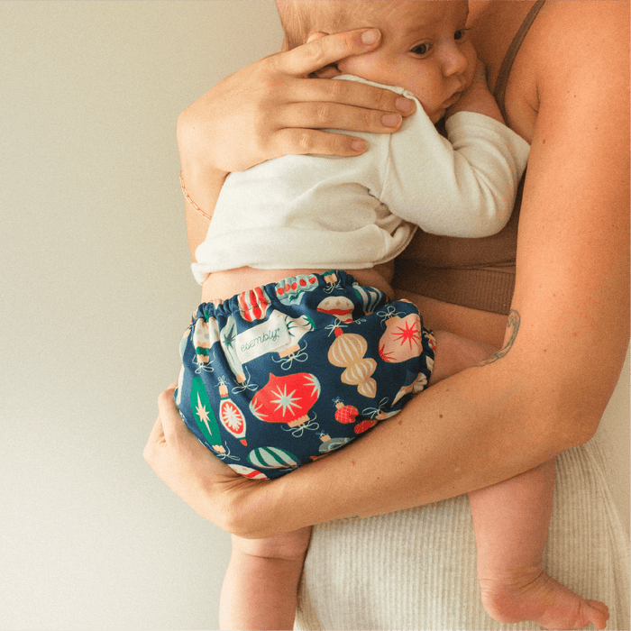 Woman holding baby wearing an Esembly Outer in Ornaments (a dark blue outer with illustrations of colorful, vintage ornaments)