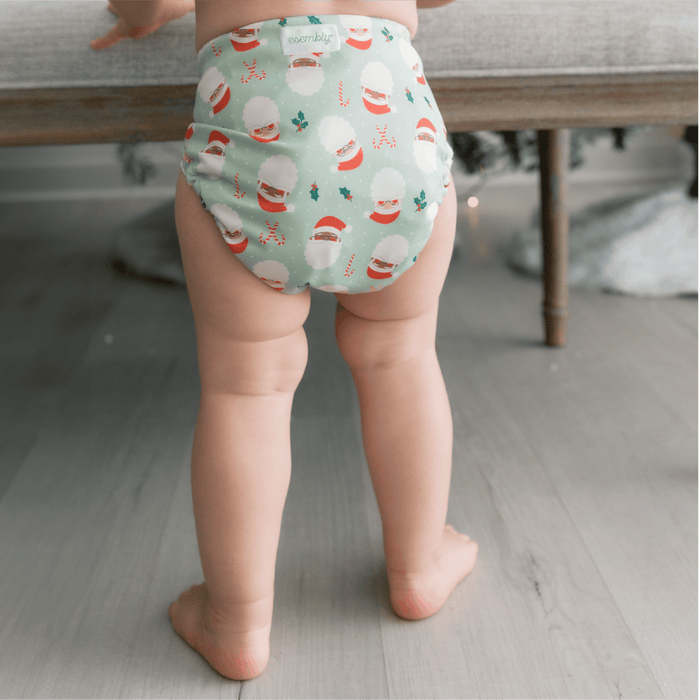 Baby standing wearing a Santas Outer (a mint green Outer with illustrated Santa heads, mistletoe, and candy canes)