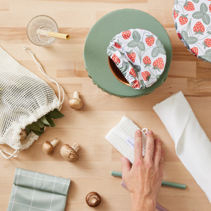 Image of our home collection that has our Produce Savers with mushrooms and greenery coming out the top, our Snack and Sammy pouch in lattice (green with an outlined check pattern), Sippers in green, yellow (in a glass), and purple, Paperless Towels, and Bowl Caps in Strawberries (a white background with red strawberries and light green leaves) and mid-tone green