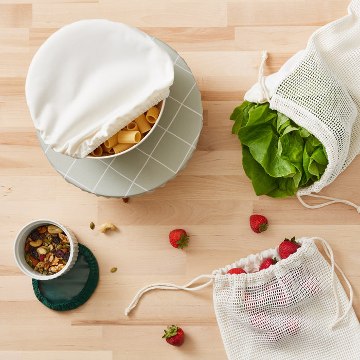 A bowl topped with a bowl cap in white with dry pasta peeking out of the bowl sitting on top of a bowl with a Lattice (a light green background with white lined grid pattern) bowl cap on top sitting on a butcher block next to a cup of assorted nuts with a dark green bowl cap, and produce savers with strawberries and spinach coming out of the bag