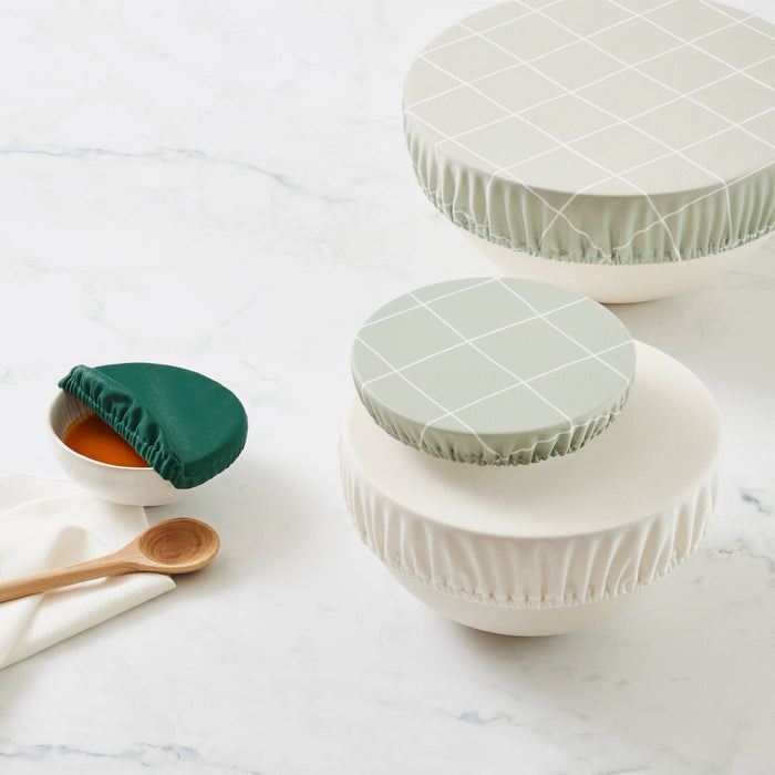 Four bowls with bowl covers (two in Lattice -- a light green background with white lined grid pattern) one in off-white, and one in dark green. The bowl with the dark green bowl cap has pasta sauce and is next to a wooden spoon and on top of a paperless towel