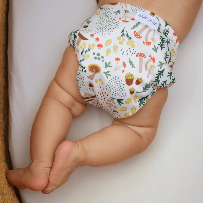 A baby laying on their stomach wearing a Fantastic Fungi Outer (an off-white background with colorful mushroom, trees, and leaves illustrations)