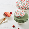 Four bowls with bowl covers (Strawberries (a white background with red strawberries and light green leaves), mid-tone green, and reddish brown. The bowl with the light green bowl cap has strawberries in and next to it and is next to a wooden spoon and on top of a paperless towel