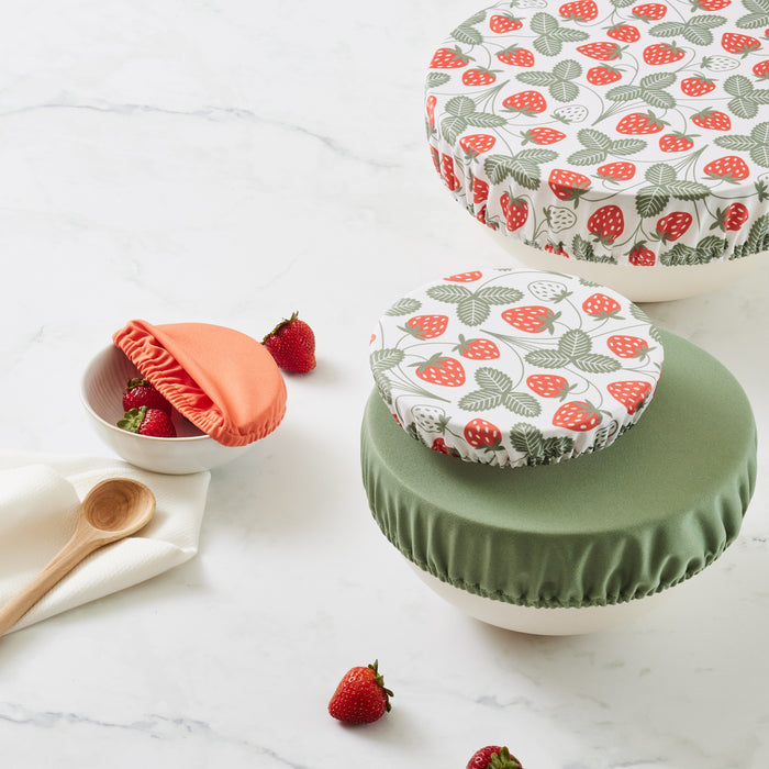 Four bowls with bowl covers (Strawberries (a white background with red strawberries and light green leaves), mid-tone green, and reddish brown. The bowl with the light green bowl cap has strawberries in and next to it and is next to a wooden spoon and on top of a paperless towel
