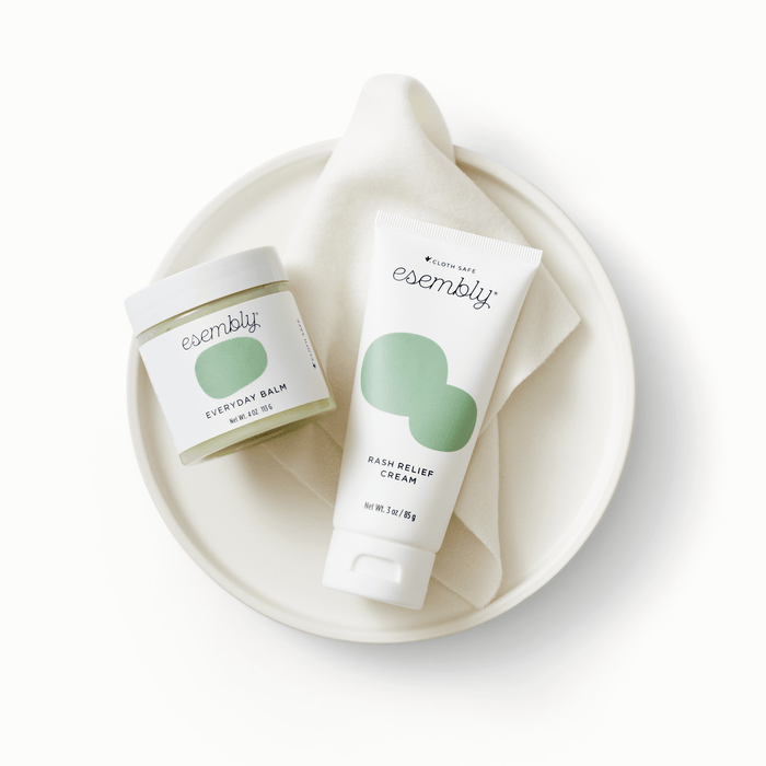 Esembly's Everyday Balm canister and Rash Relief Cream tube sitting on top of a white towel on top of a white plate