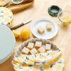 Lemon bars on a table with a lemon-themed Bowl Caps, green Bowl Caps, and lemons.
