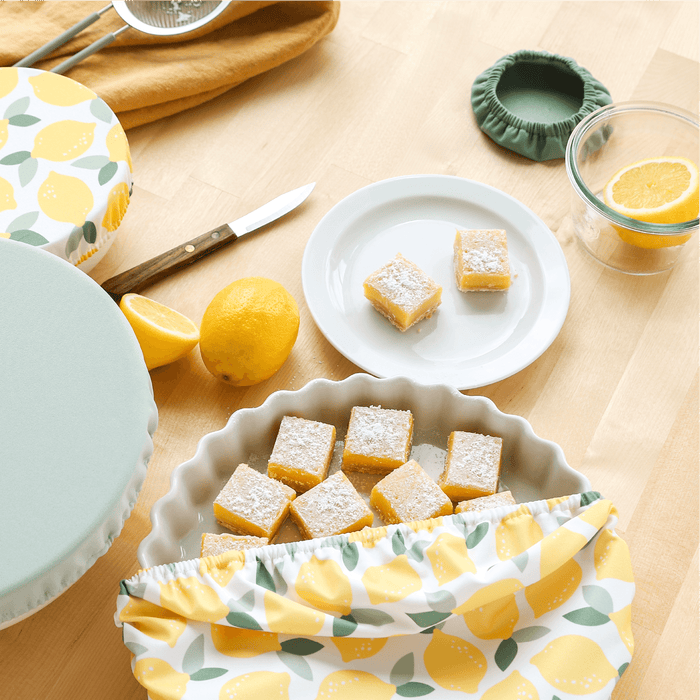 Lemon bars on a table with a lemon-themed Bowl Caps, green Bowl Caps, and lemons.