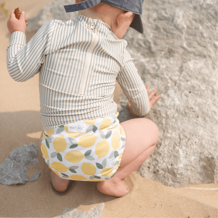 Child wearing a striped shirt and a diaper with lemon pattern on a sandy beach.