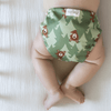 A baby laying on their stomach wearing an Esembly Cloth Diaper Outer in Peek-A-Bear (mid-tone and light green trees in the background with illustrated brown bears peeking out)