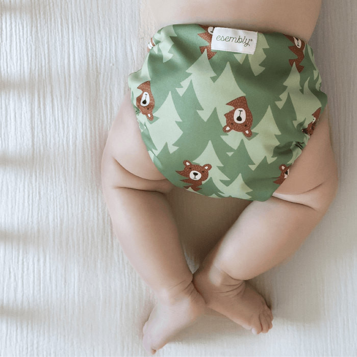 A baby laying on their stomach wearing an Esembly Cloth Diaper Outer in Peek-A-Bear (mid-tone and light green trees in the background with illustrated brown bears peeking out)