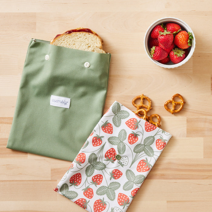 A reusable sandwich bag in mid-tone green with a sandwich peeking out of the top next to a reusable snack bag in Strawberries (a white background with red strawberries and light green leaves) with pretzels coming out of the top next to a small cup filled with strawberries and everything sitting on a butcher block table top