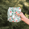 A hand holding a Santas Outer (a mint green Outer with illustrated Santa heads, mistletoe, and candy canes) in front of a pine tree
