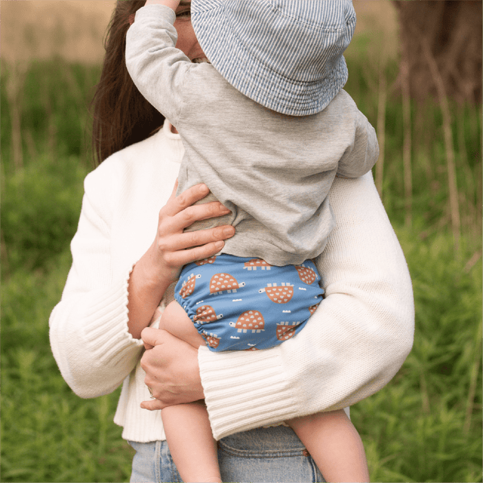 Person holding a child wearing a Turtle patterned diaper in a grassy outdoor setting