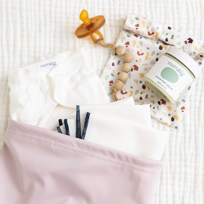 Front view of a waterproof bags with toggle close top in light purple with a white cotton diaper, white cloth wipes, a pacifier and a plastic jar of diaper balm