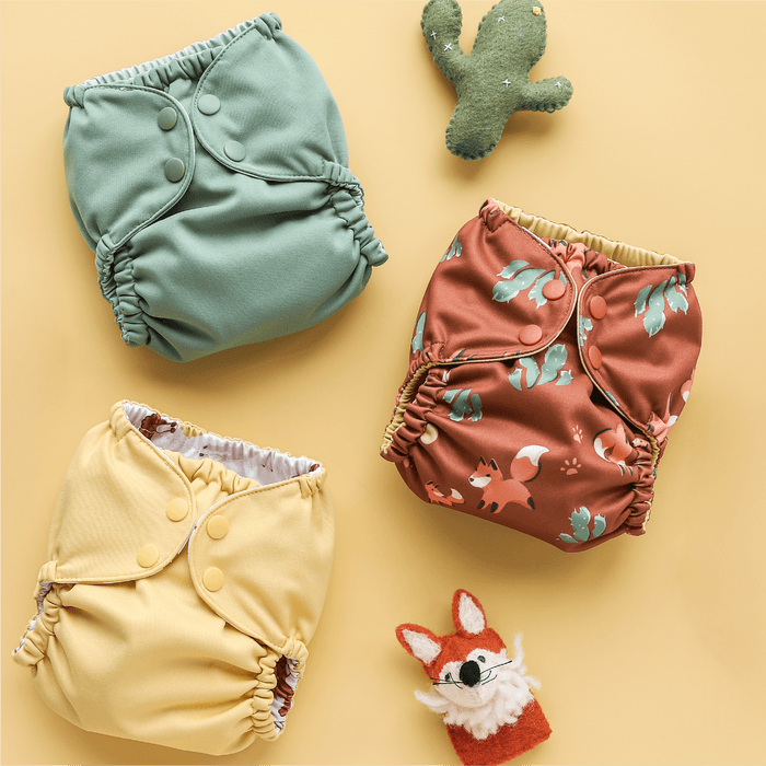 Three colorful baby diapers on a yellow background with small fox and cactus felt toys.