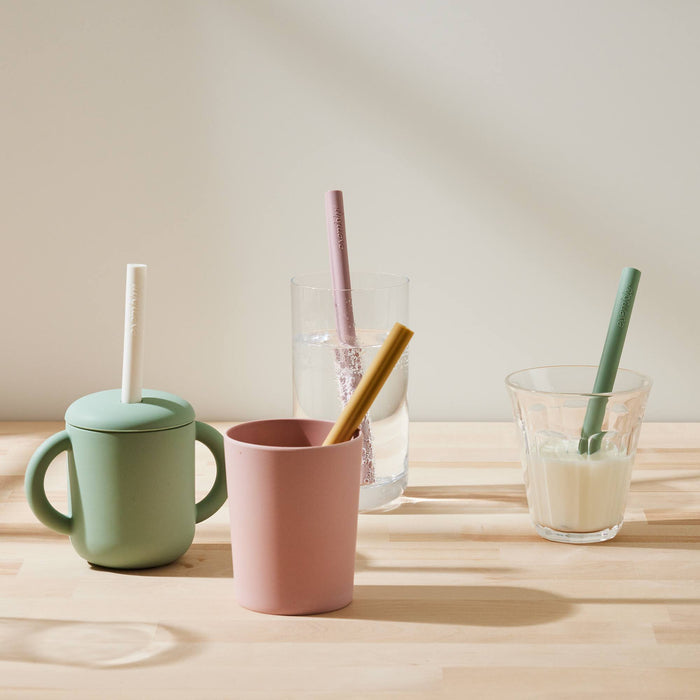 A mint green cup with handles with a white silicone straw coming out the top, a light pink plastic cup with a muted yellow straw coming out the top, a clear glass with water in it and a pink silicone straw coming out the top, and a glass with milk and a green silicone straw in it. All sitting on a butcher block counter top