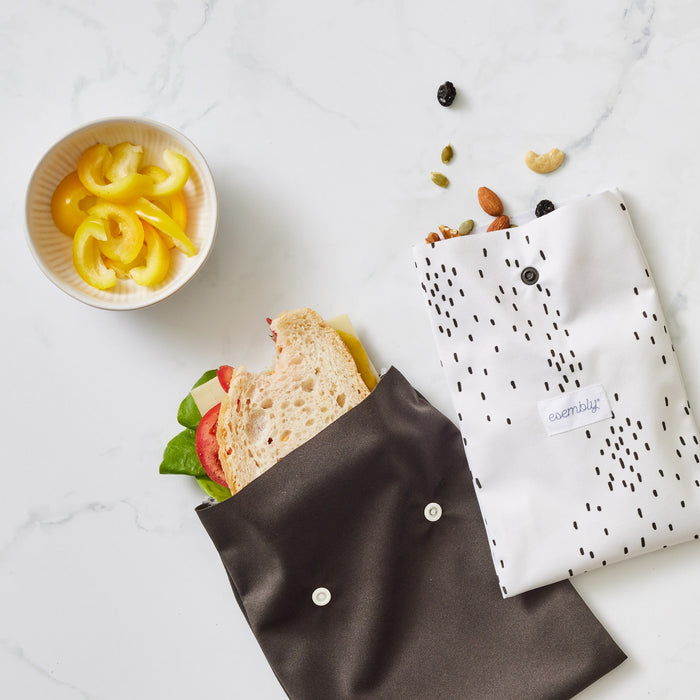 A black reusable sandwich bag with a sandwich coming out of it and a reusable snack bag in Apostrophe (a white background with black speckles) with mixed nuts coming out the top sitting on a marble countertop next to a small bowl with sliced yellow peppers