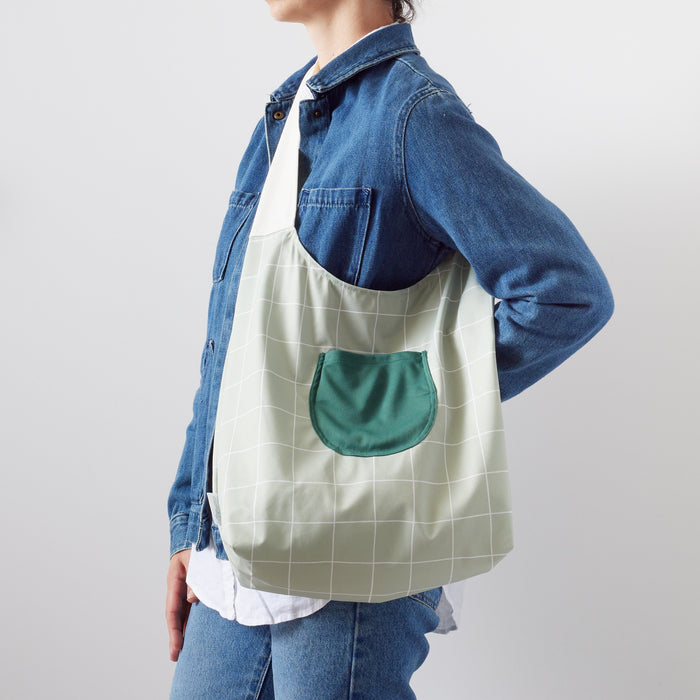 Esembly Lattice Carryall Bag (a Lattice (a light green background with white lined grid pattern) body, dark green pocket, and an off-white strap) on a woman wearing a denim jacket, jeans, and a white button down