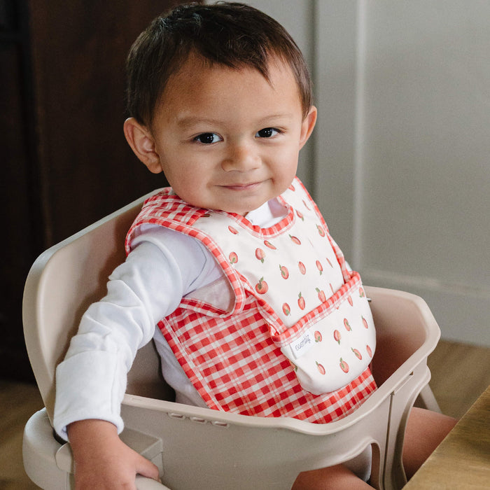 Baby wearing an Esembly bib with apples and red plaid on it. The baby is sitting in a high chair in a kitchen.
