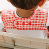 Close-up back view of a child wearing a red and white plaid bib over a white shirt, seated in a cream-colored high chair.