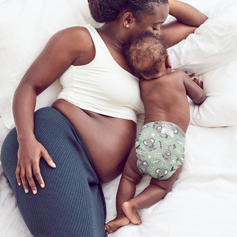 Back-view of baby wearing an Elephants Outer (a light green background with light grey elephants and white and black tree limbs and white sporadic polkadots ), crawling next to a woman wearing a grayish blue skirt with a white bra, laying on her side on white bed and pillows