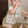 A baby standing at a high chair wearing a cream bib with a green-trimmed front pocket and a tractor print.