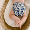 Baby laying in a wicker changing basket wearing an Outer with a white background with dark navy blue and light blue blueberries with light green leaves