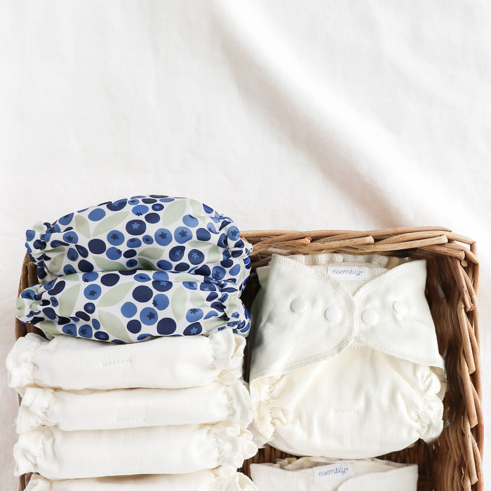 Two Esembly x Winter Water Factory Outers in Blueberries (blueberries in three shades of blue and light green leaves) and Esembly Inners in a brown wicker basket sitting on a crisp white sheet