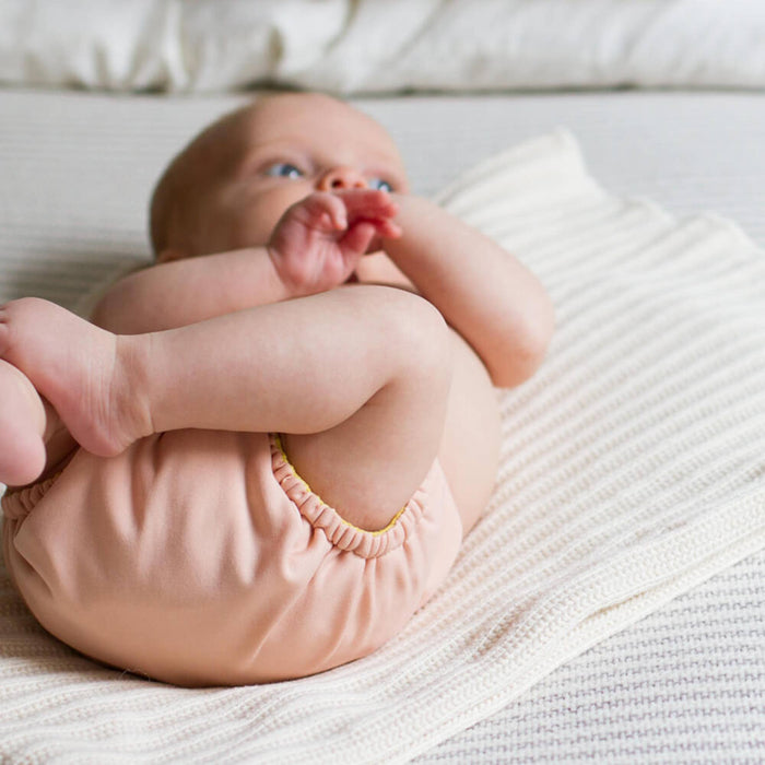 Baby laying on a white blanket on a bed wearing an Outer that is a light pink color