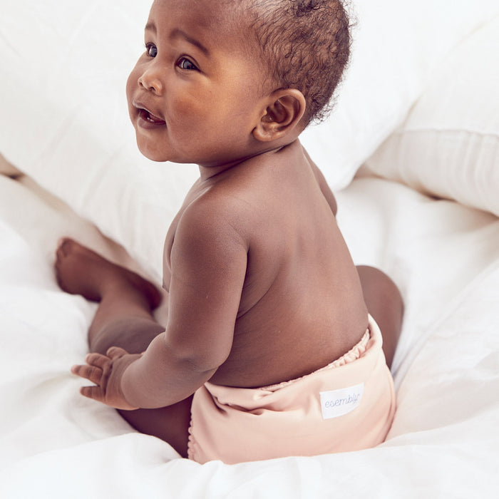 Baby sitting on fluffy white blanket on bed wearing an Outer in a light pink color