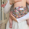 A baby wearing an Esembly Outer in Botanic Garden (colorful florals hand painted by illustrator Sara Boccaccini Meadows) being held by their mom who is wearing white