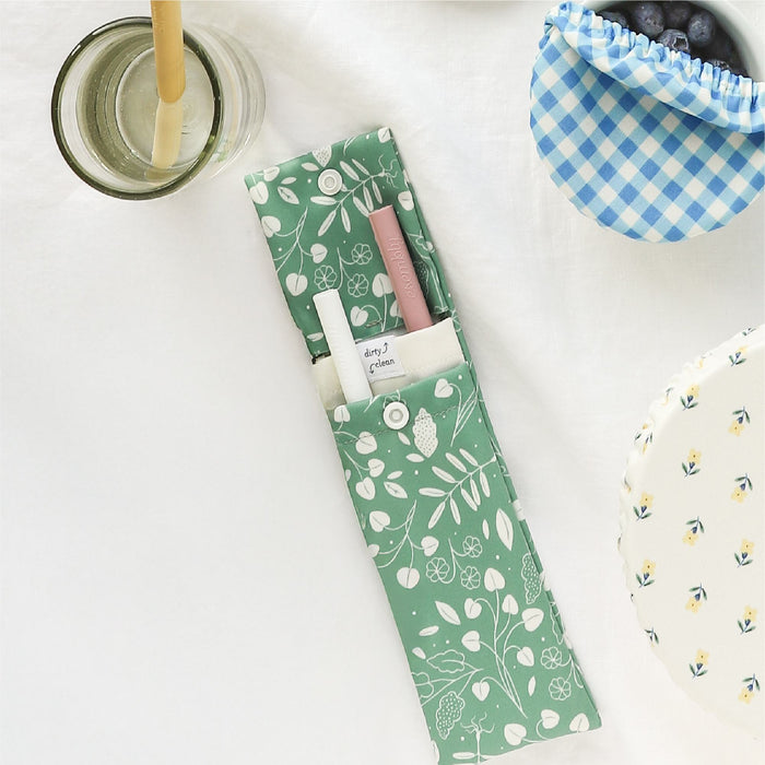 A green patterned fabric pouch containing eco-friendly silicone straws, sits on a white surface alongside. Nearby, there's a silicone straw in a water glass, a bowl with blueberries covered by a blue gingham bowl cap, and a bowl covered by a Buds floral and white background bowl cap.