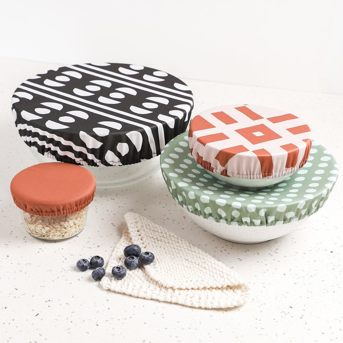Image of four bowl caps in varying shapes on bowls on a terrazzo countertop next to blueberries and a dish cloth. One bowl cap in Circuit (a black and white pattern with a half circle and organic circle pattern), a bowl cap in Stellar (a mid-tone green background with light blue organic shaped dots), a bowl cap in Theory (a reddish brown square and line geometric pattern on a light purple background), and a solid reddish brown bowl cap.
