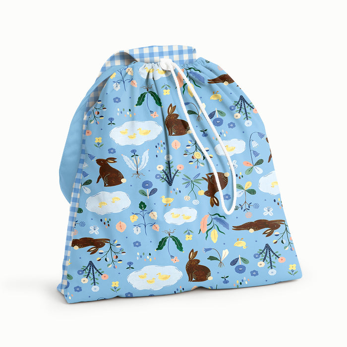 Front View of an Esembly Day Bag with Bunnies on the front (bunnies, flowers, and ducks illustrated by Flora Waycott on a mid-tone blue background), blue gingham on the flap and back, and a mid-tone blue strap