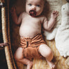 A baby laying in a wicker basket next to a stuffed animal wearing an Outer in a medium brown color