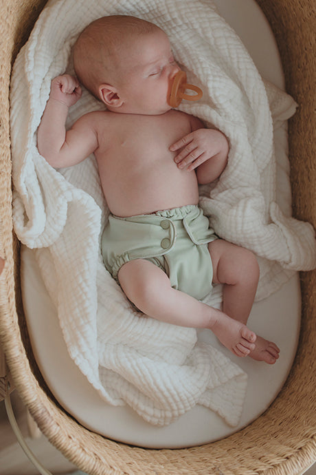 Baby in a woven bassinet wearing an Esembly Outer in Sea Glass (a light green color)