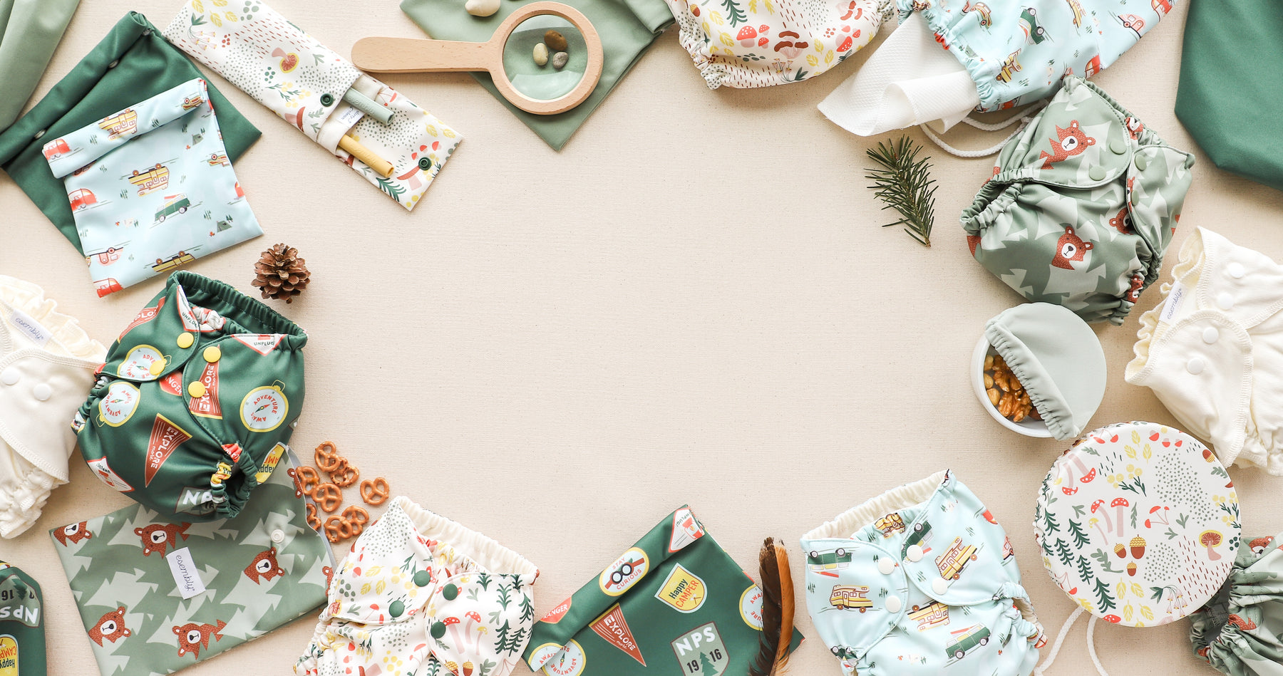 Reusable cloth diapers in Esembly's National Parks patterns are arranged in a circle on a white surface. Accessories like a magnifying glass, cloth wipes, and a small branch are also included in the arrangement.