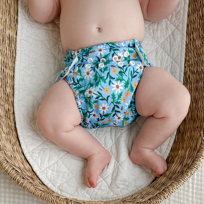 Baby laying on a white mat in a wicker basket wearing an Outer with a light blue background with white and yellow flowers with light green and dark green leaves