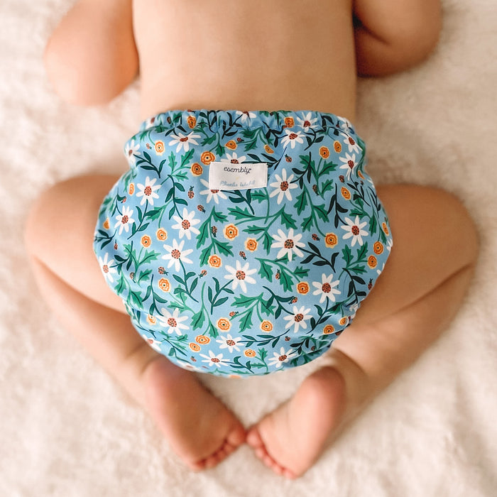 Baby laying on a white blanket wearing an Outer with a light blue background with white and yellow flowers with light green and dark green leaves