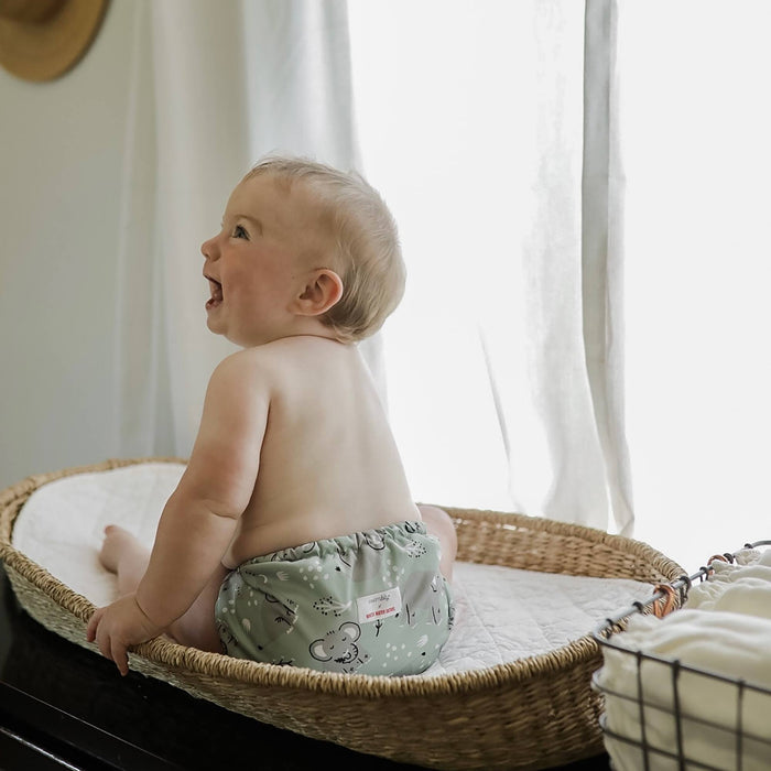 A baby sitting on a wicker changing pad wearing an Outer with a light green background with light grey elephants and white and black tree limbs and white sporadic polkadots 