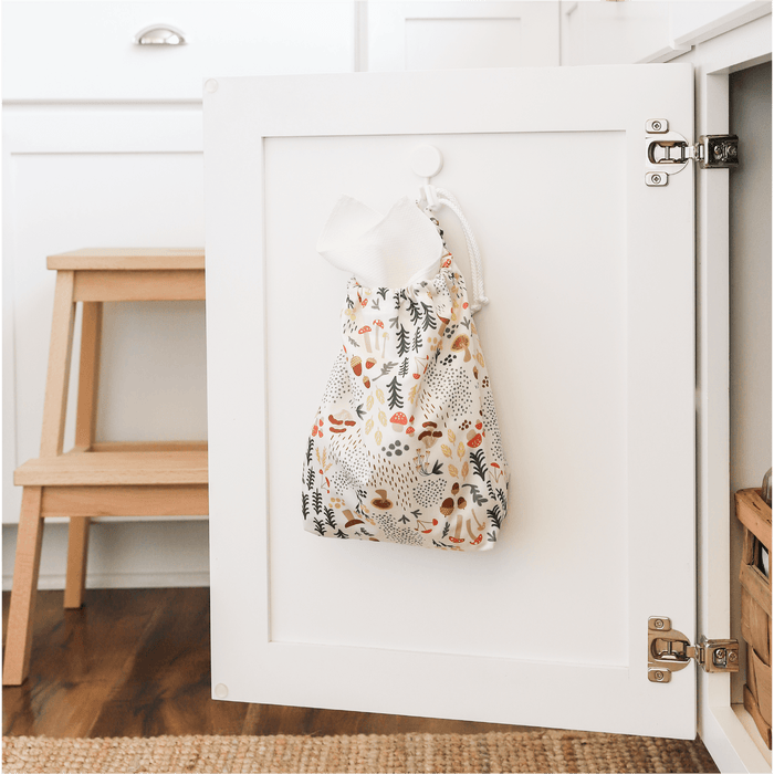 Paperless Towels in Fantastic Fungi (an off-white background with colorful mushroom, trees, and leaves illustrations) hanging inside of a white kitchen cabinet