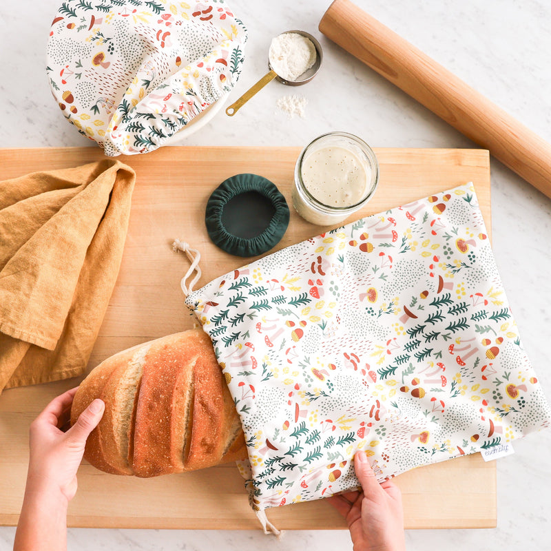 Hand pulling a large loaf of bread out of the Sourdough Set in Fungi (an off-white background with colorful mushroom, trees, and leaves illustrations)