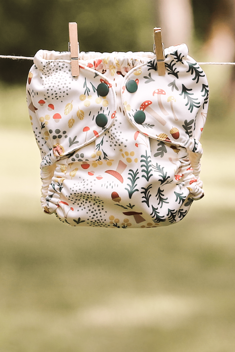 A Fantastic Fungi Outer (an off-white background with colorful mushroom, trees, and leaves illustrations) hanging on a laundry line with clothing pins