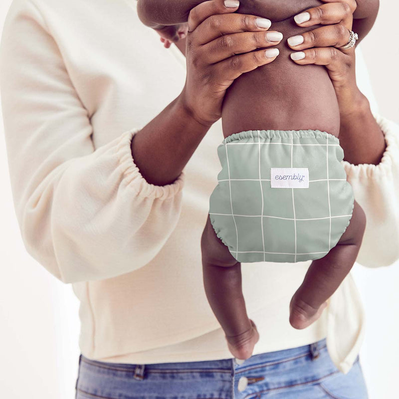 Mom in a cream colored shirt and jeans holding up her baby wearing an Esembly Outer in Lattice (a mid-tone green color with a large off-white outlined check pattern)